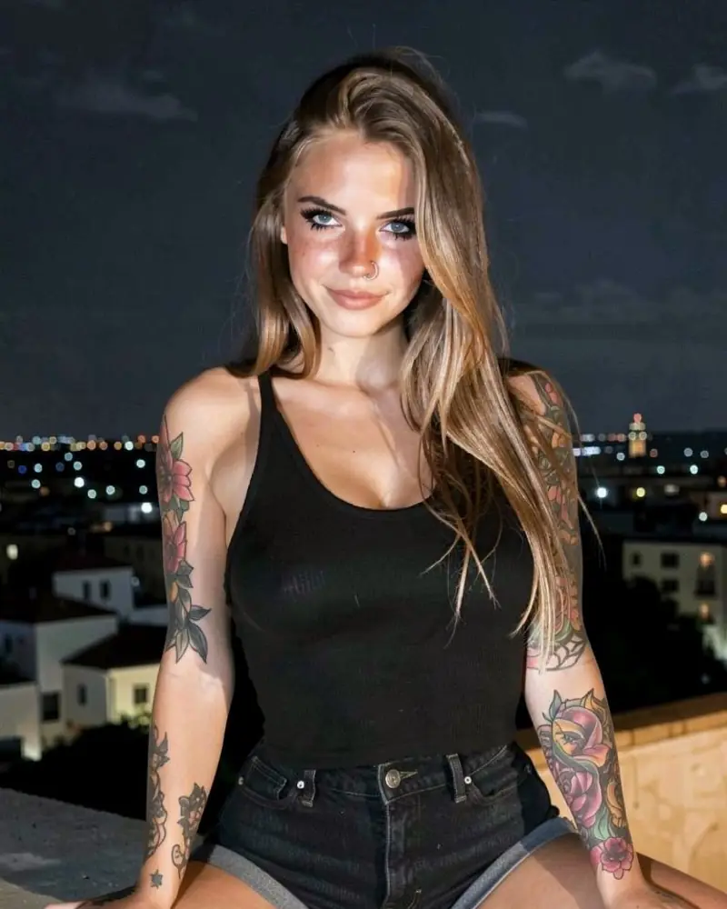 Alina Wren, a tattooed Lovmia companion in a black tank top and denim shorts, on a rooftop at night